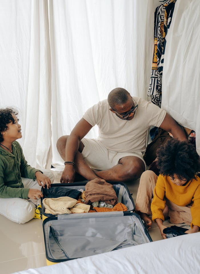 From above of young African American man sitting on floor and packing suitcase with little daughters